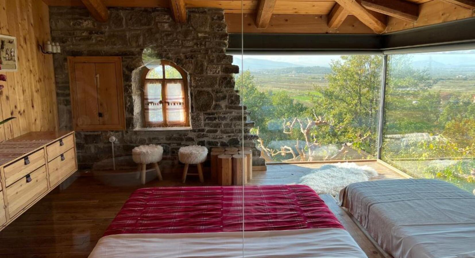 Albanian mountain guesthouse room — stone walls, timber beams, red blanket and valley view through glass